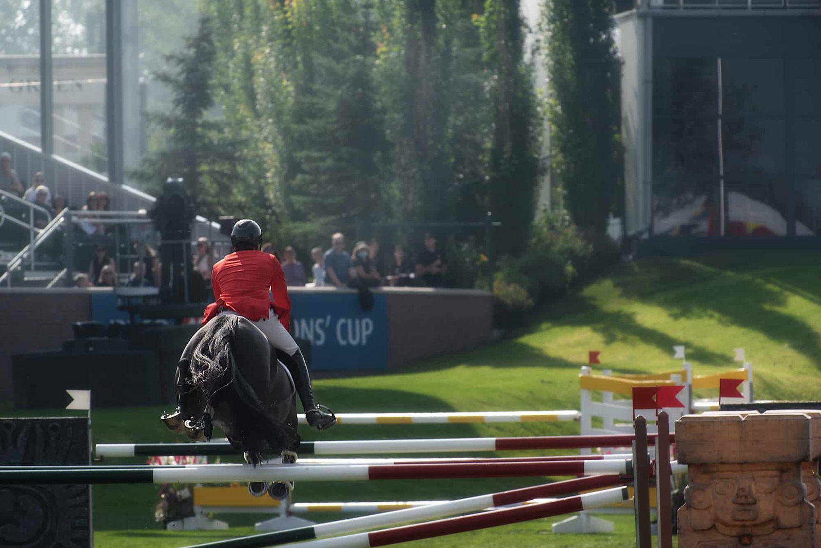 A horse and rider combination competing in the iconic international ring at Spruce Meadows in Calgary, Alberta. The rider, dressed in formal showjumping attire, expertly guides the horse over a jump adorned with colorful poles and sponsor banners. The lush green grass of the arena contrasts with the vibrant flowers and the grandstands filled with spectators in the background, capturing the prestige and energy of the event.